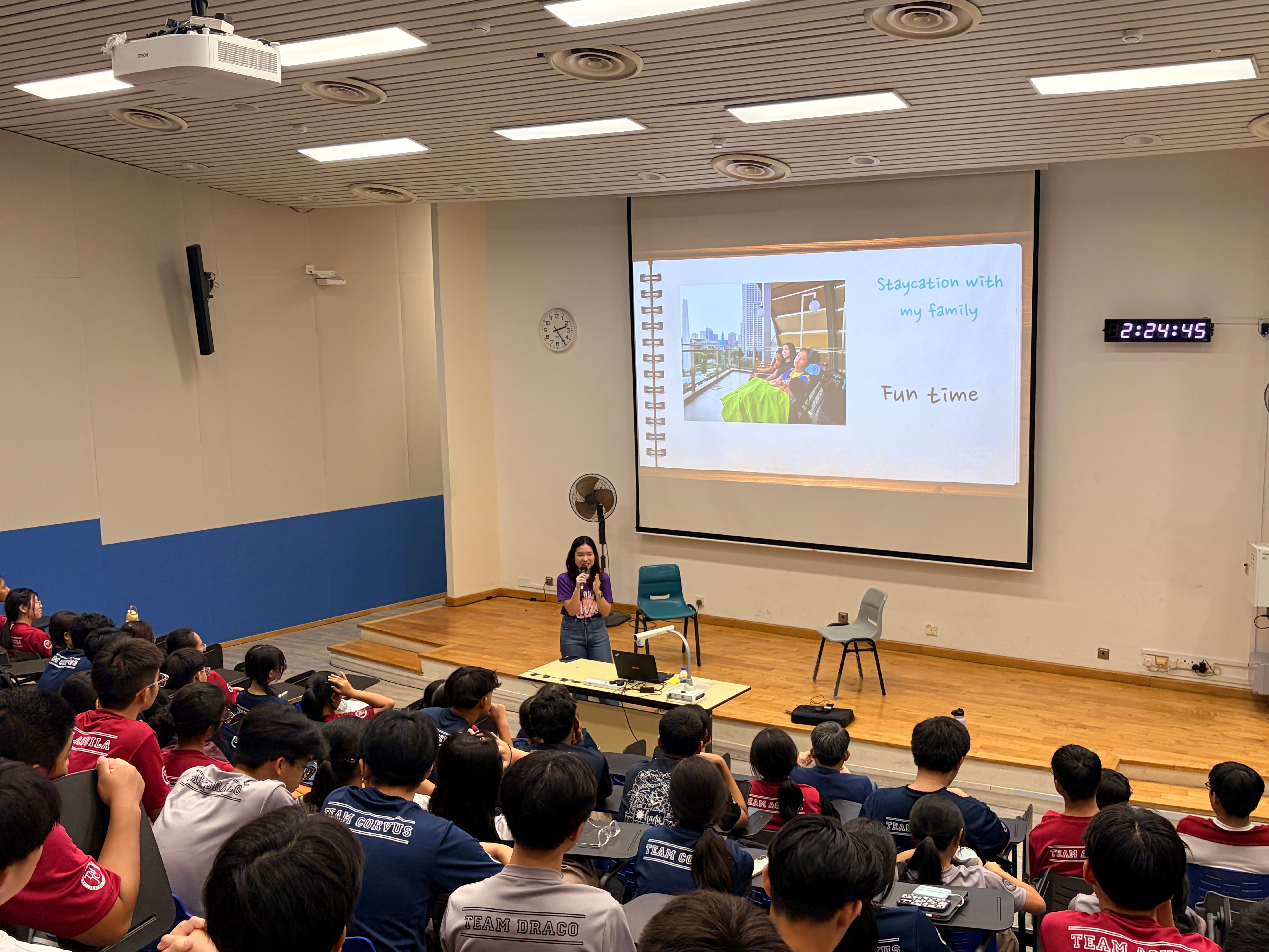 A young woman speaks into a microphone before a packed lecture theatre, with a slide showing "Staycation with my family – Fun time" projected behind her.